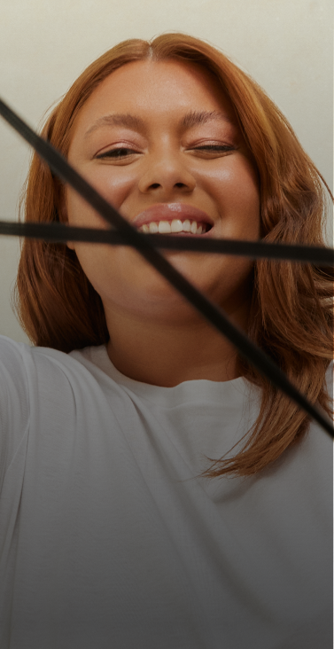 Uma mulher de pele morena e cabelos ondulados ruivos sorri enquanto olha para a câmera e faz o símbolo do logo do next com um elástico nas mãos, que lembra o símbolo do infinito.