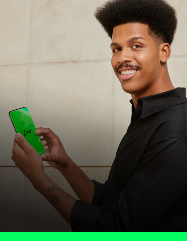 Vemos um homem negro, de cabelo crespo e bigode, sorrindo. Ele está vestindo uma camisa na cor preta, posicionado de lado, com a cabeça virada para olhar para câmera. Em suas mãos, vemos um celular com a tela verde, mostrando o logo do next.