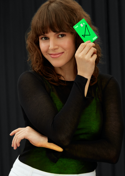 Vemos uma mulher de cabelos até o ombro e ondulados, com franja, olhando para a câmera e sorrindo, ela também segura ao lado do rosto o cartão verde do next.