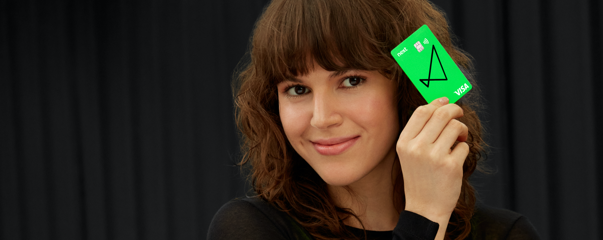 Vemos uma mulher de cabelos até o ombro e ondulados, com franja, olhando para a câmera e sorrindo, ela também segura ao lado do rosto o cartão verde do next.